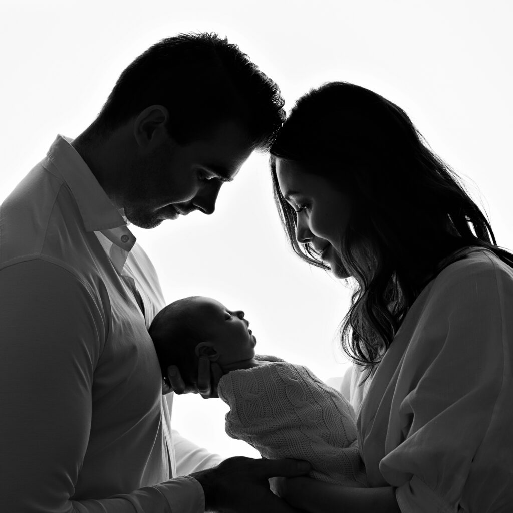 Silhouette of parents gazing lovingly at their newborn baby, captured in black and white, highlighting the tender bond and affection within the family.